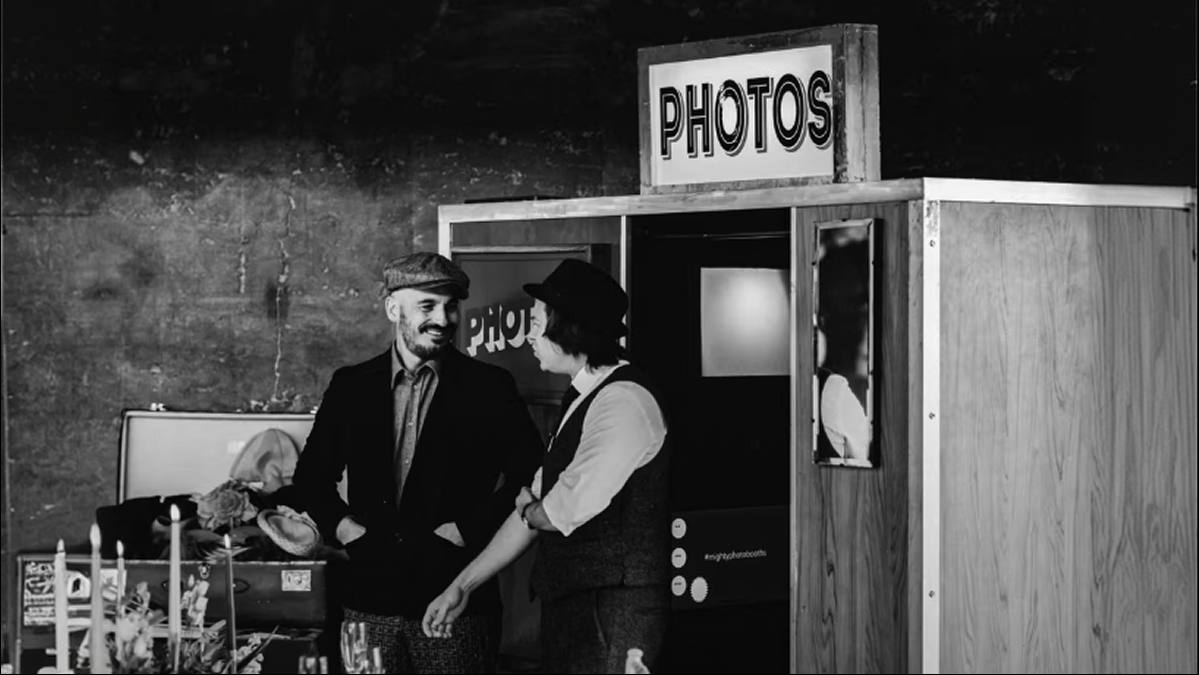 Black-and-white photo of two guests in hats talking beside a timber photo booth at an event