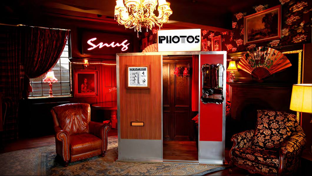 Timber and red photo booth with a PHOTOS sign installed in a chandelier-lit red lounge with a Snug neon sign